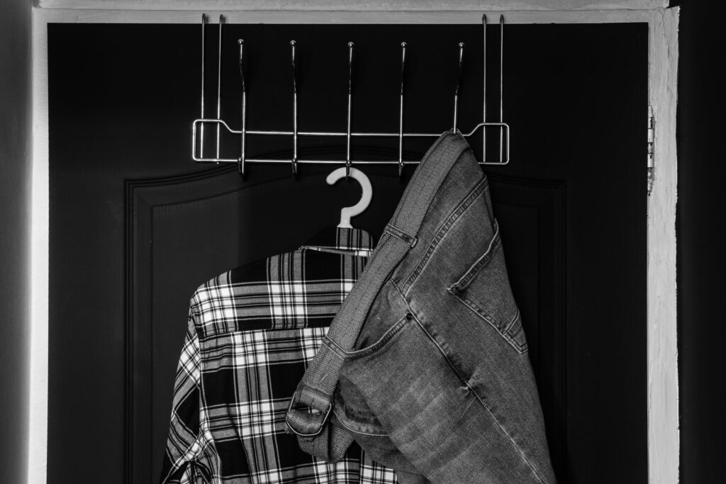 Black and white photo of a metal over-the-door hook rack holding a plaid shirt and jeans, with a question mark-shaped hanger.