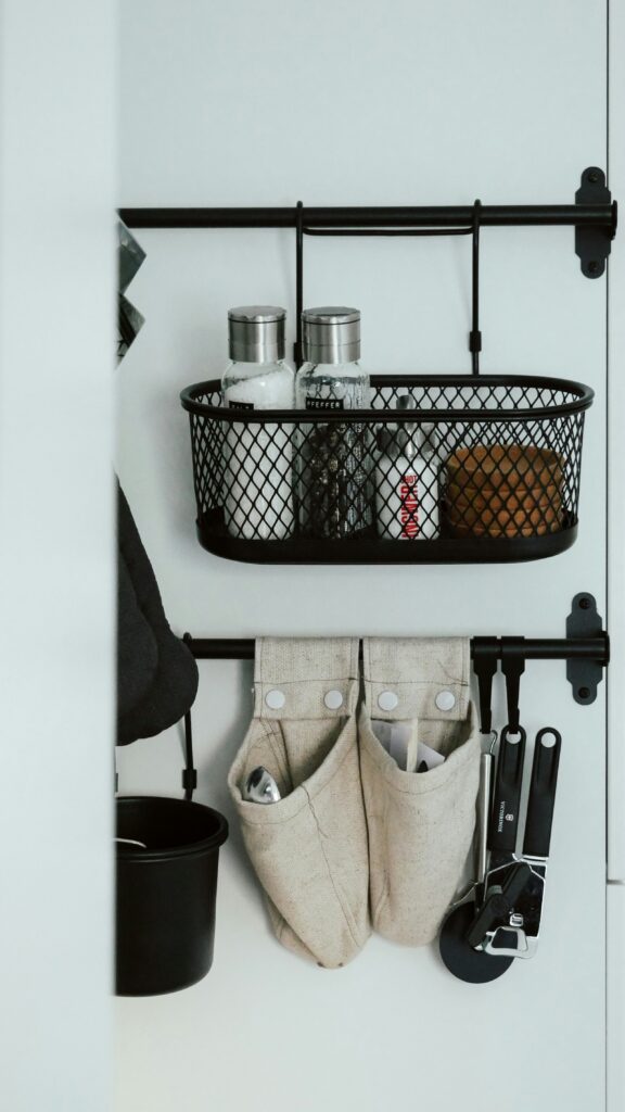 Black mesh storage basket hanging on a white door, holding spices and kitchen utensils.