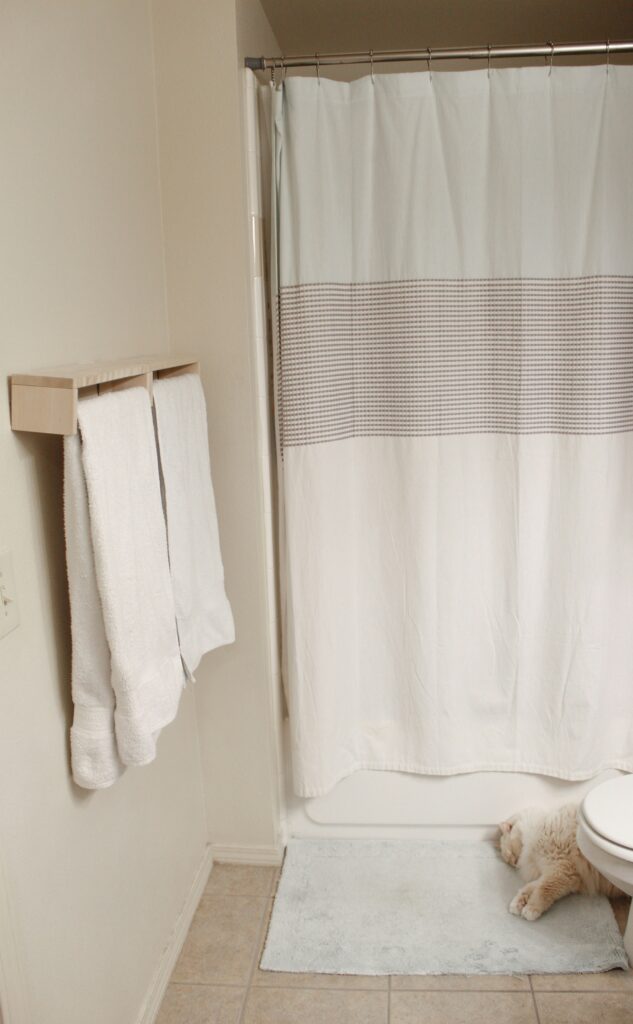 Floating wooden towel rack mounted on beige wall next to shower curtain, holding two white towels