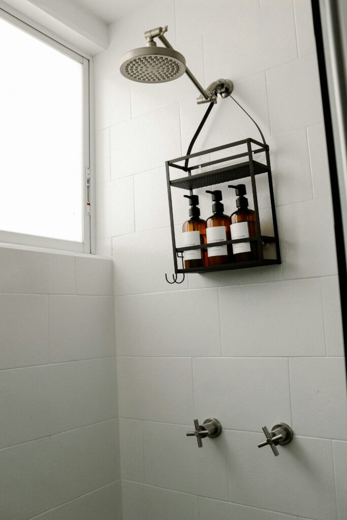 Black metal hanging shower organizer suspended from showerhead pipe, holding three amber pump bottles