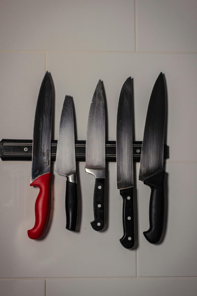 Black stainless steel magnetic knife strip holding professional kitchen knives with black and red handles on a tiled wall